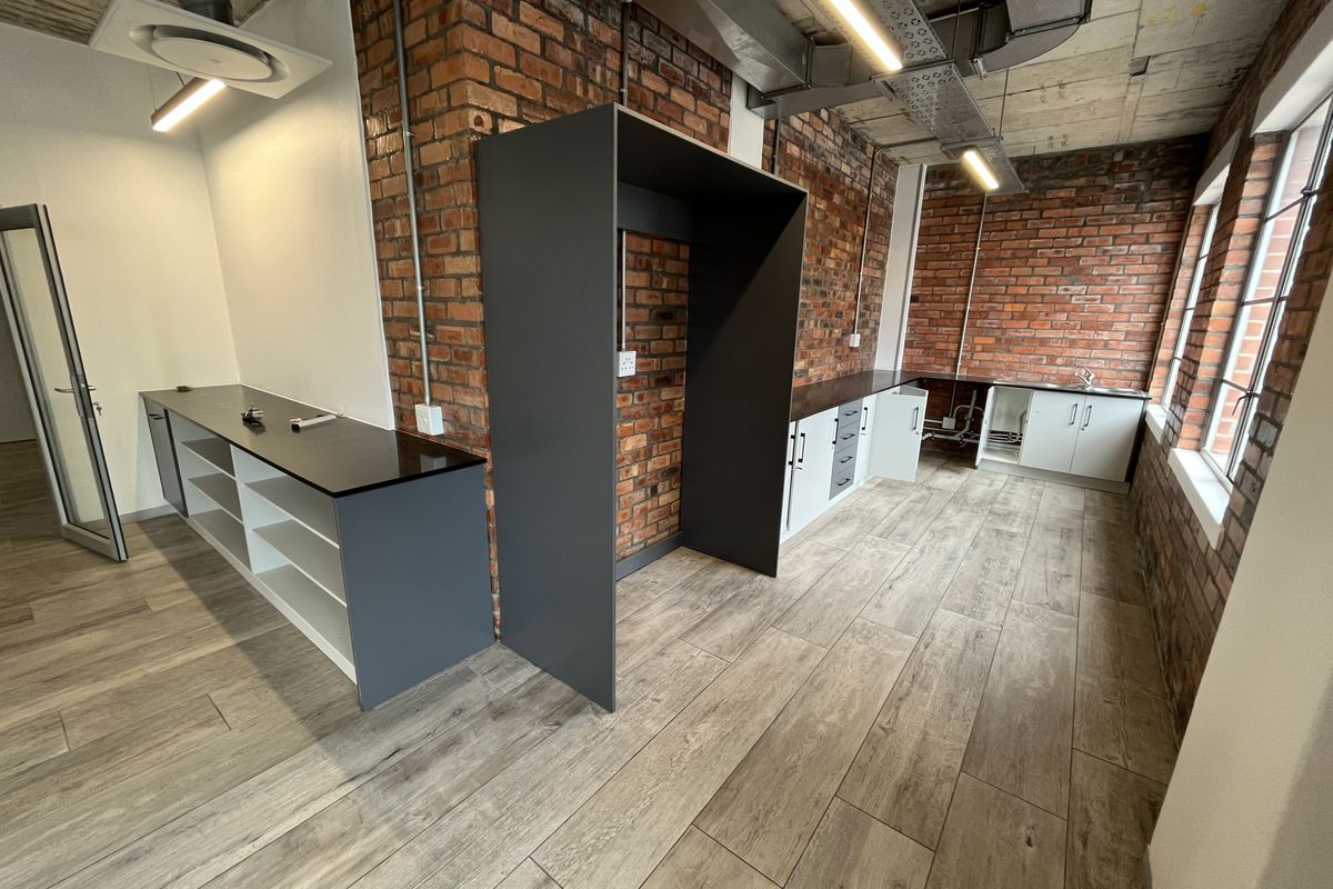 Modern office kitchenette featuring exposed brick walls, industrial-style ceiling, wood-look tile flooring, and sleek built-in cabinetry with integrated workspace.