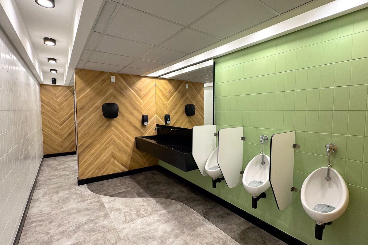 Modern office restroom featuring wall-mounted urinals with privacy partitions, a sleek black trough sink, and contemporary green and wood-look wall tiling.