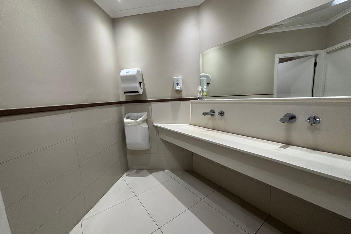 A clean and modern office restroom featuring a long vanity counter, wall-mounted fixtures, and tiled flooring.