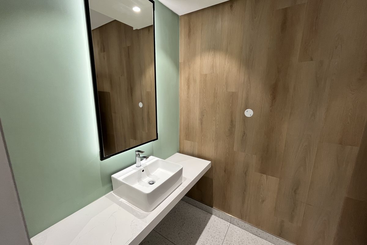 Modern office restroom featuring a sleek vessel sink, stone countertop, backlit mirror, and contemporary wood-look wall paneling.