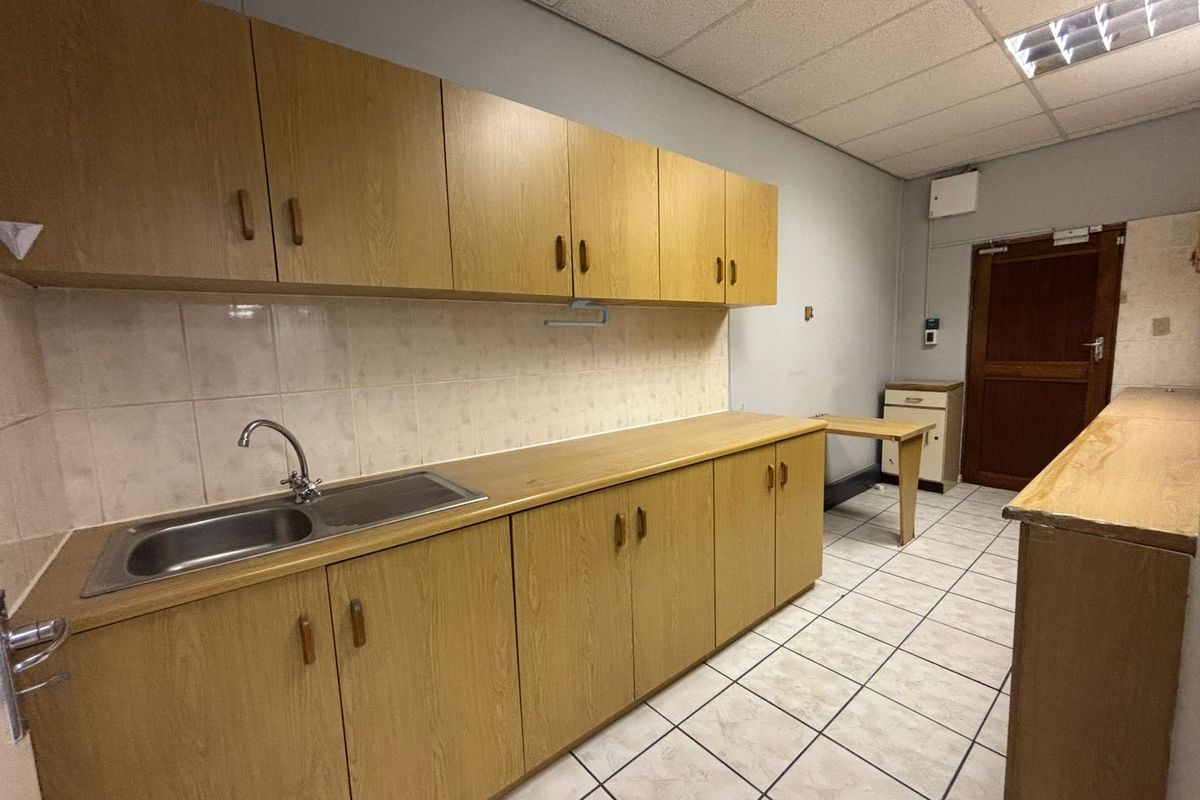 A functional kitchenette area featuring wood-laminate cabinetry, a stainless steel sink, tiled splashbacks, and a suspended ceiling with integrated lighting.