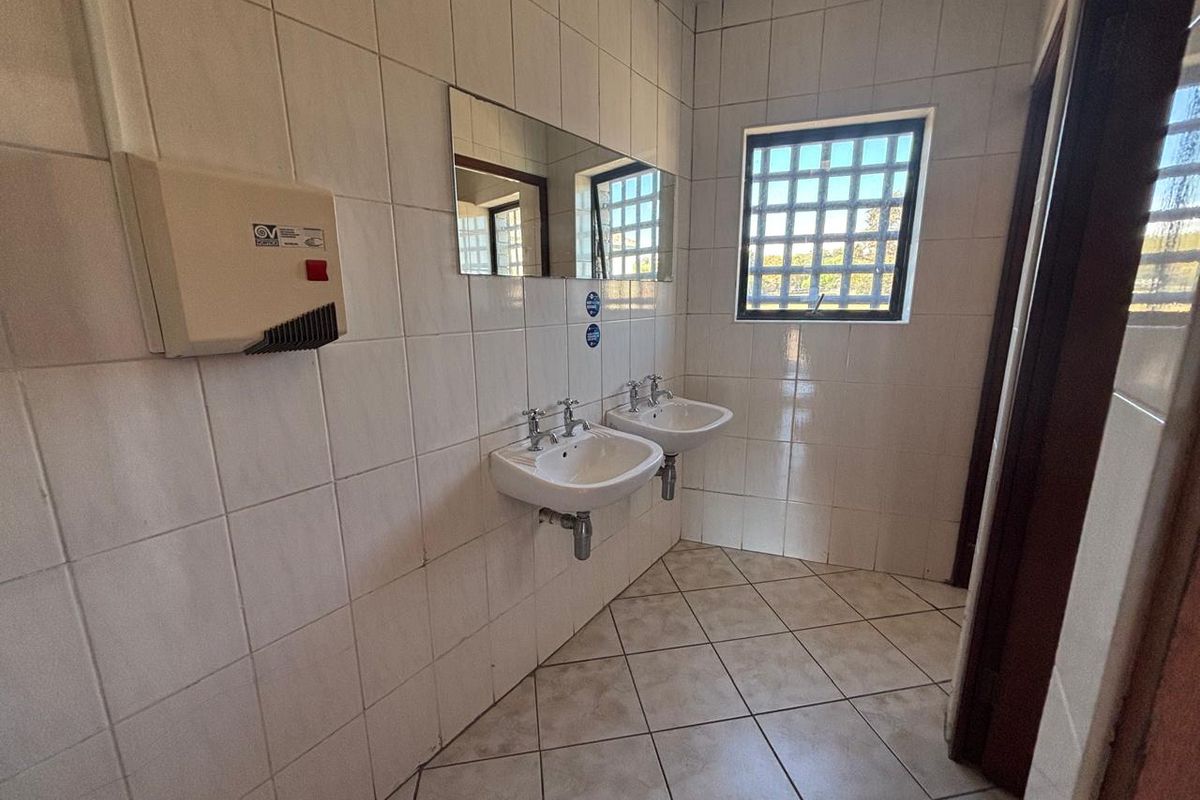 A functional office restroom featuring dual wall-mounted ceramic basins, tiled walls, and a security-grille window.
