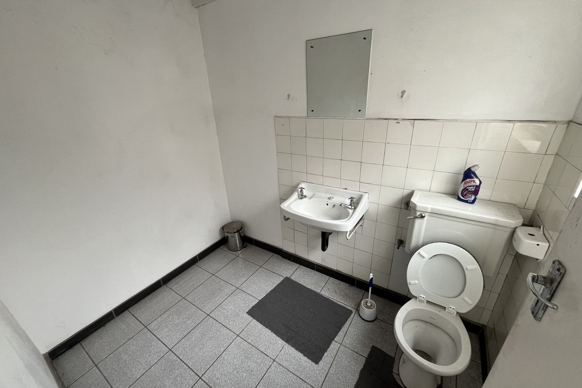 A functional office restroom featuring a wall-mounted basin, standard toilet, and tiled flooring, requiring cosmetic updates and maintenance.