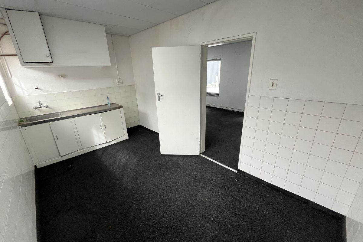 A small office kitchenette area featuring basic cabinetry, a stainless steel sink, tiled splashbacks, and dark carpeting, leading into an adjacent office space.