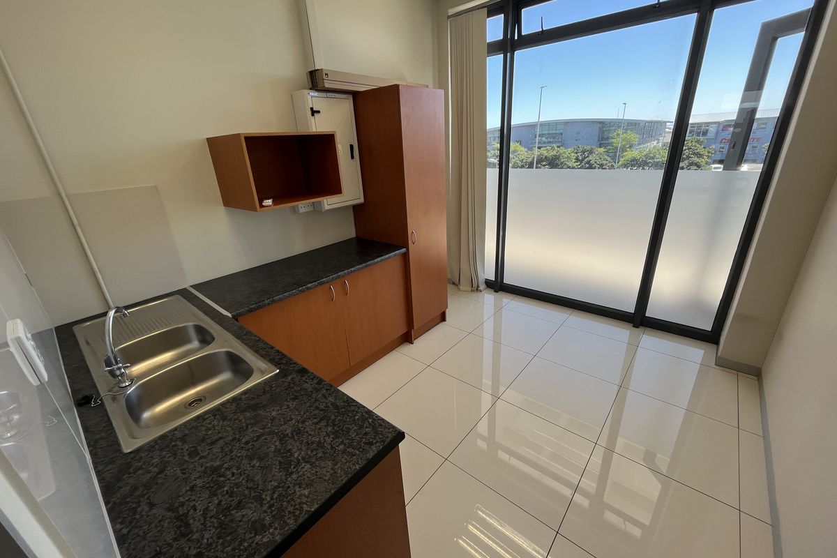 A functional kitchenette area featuring dark laminate countertops, stainless steel double sink, and built-in wooden cabinetry, set against large windows with natural light.