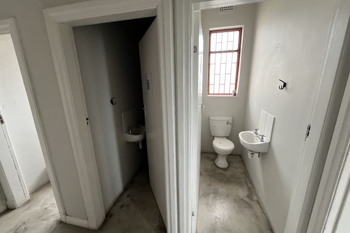 Two small, functional office restrooms featuring basic toilets, wall-mounted basins, and security-barred windows with exposed concrete flooring.