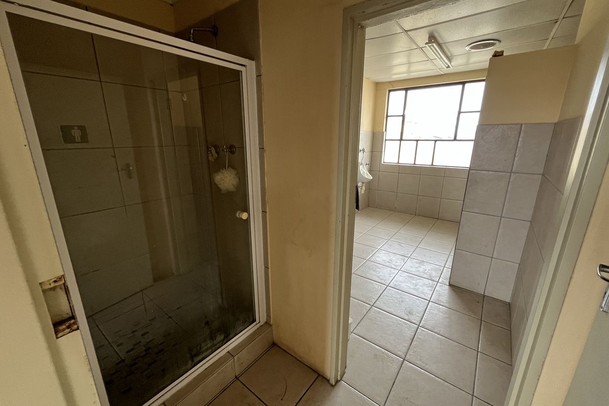 A dated office bathroom facility featuring a shower cubicle, tiled flooring, and a separate wash area with natural light.