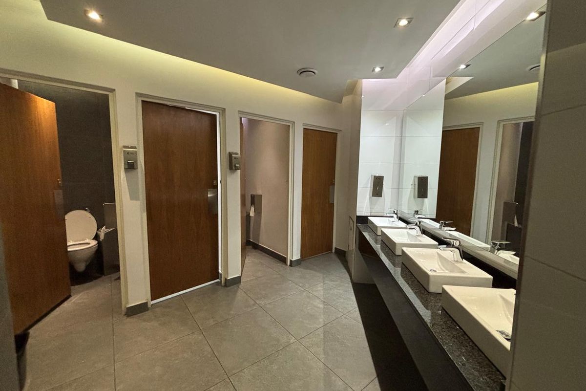 Modern office restroom featuring a triple vanity with granite countertop, large wall-to-wall mirror, recessed lighting, and multiple private toilet cubicles.