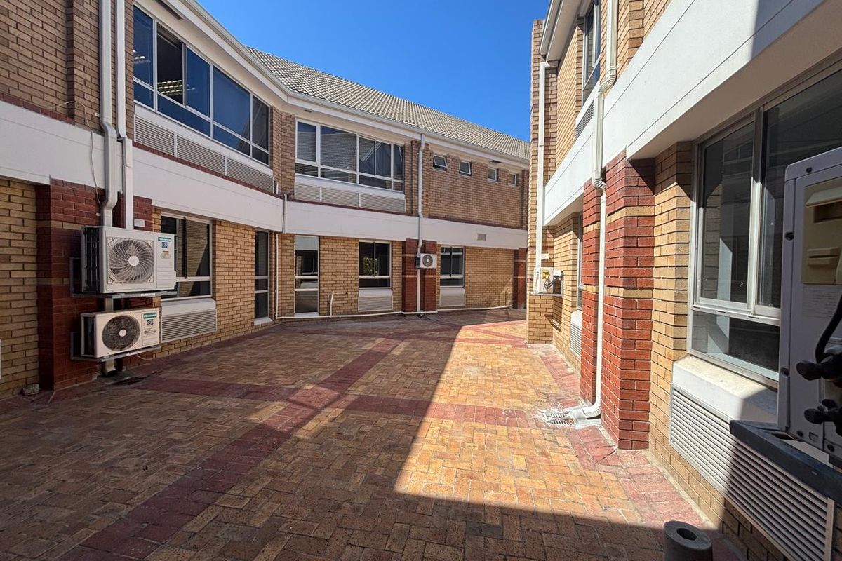 A paved exterior courtyard area between office building wings, featuring face-brick facade, aluminium window frames, and wall-mounted air conditioning units.