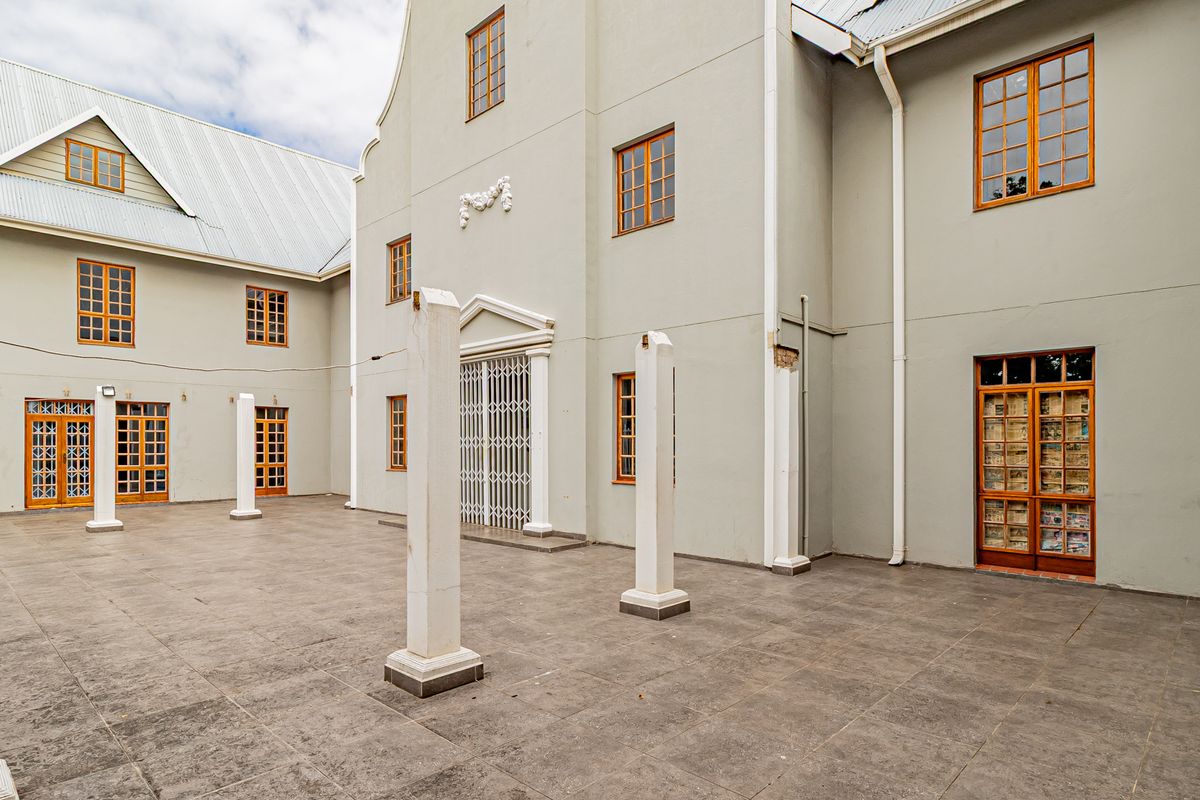 A spacious paved courtyard area featuring Cape Dutch architectural elements, multiple wooden-framed glass doors, and decorative pillars.