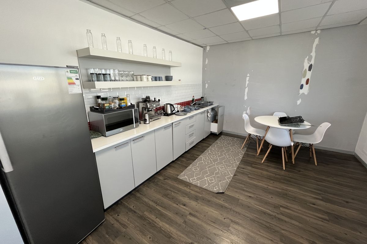 A functional office kitchenette featuring white cabinetry, floating shelves, subway tile backsplash, and wood-look flooring, currently showing signs of wall surface wear.