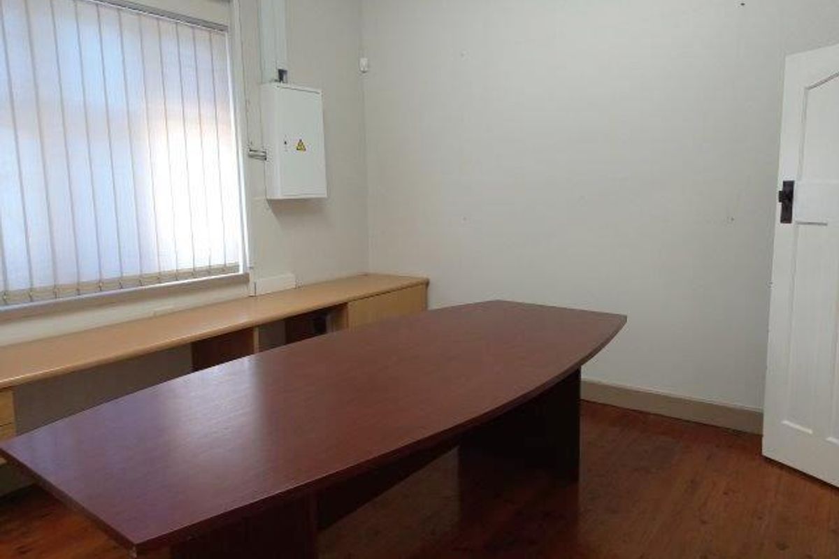 A functional office space featuring polished wooden flooring, built-in cabinetry, vertical window blinds, and a wall-mounted electrical distribution board.