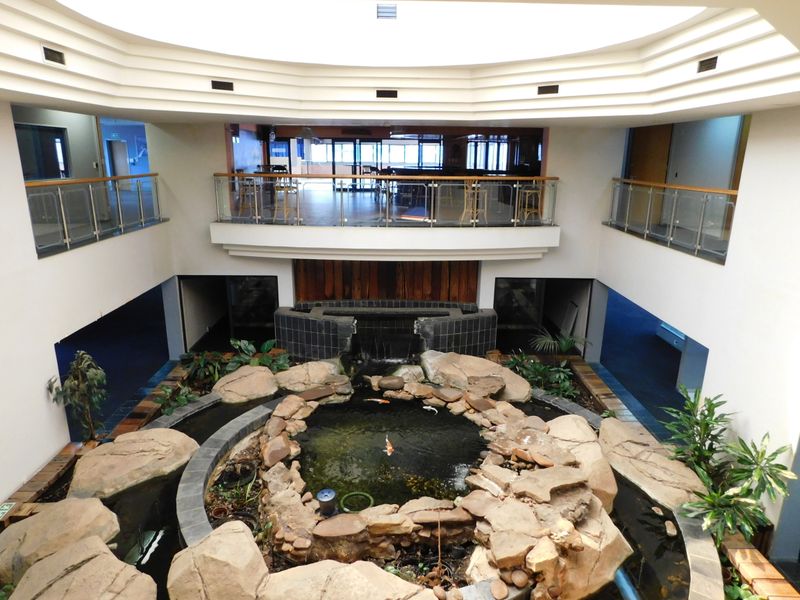 A central atrium office space featuring a decorative indoor koi pond with rock landscaping and a water feature, overlooked by a mezzanine level.