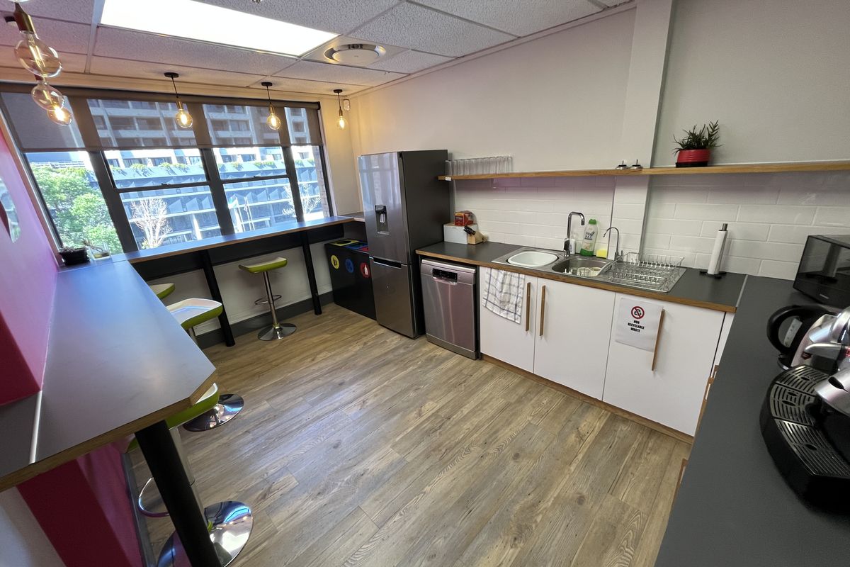 A modern office kitchenette featuring white cabinetry, a stainless steel sink, integrated dishwasher, and a breakfast bar with window views.