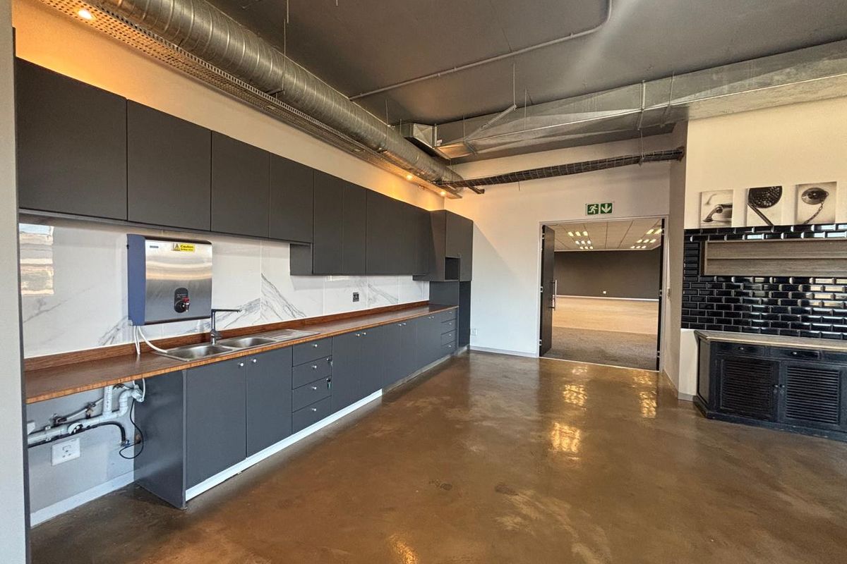 Modern office kitchenette featuring dark cabinetry, wood-look countertops, stainless steel sink, and exposed industrial-style ductwork with polished concrete flooring.