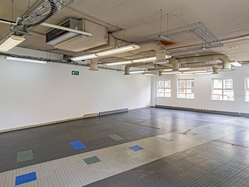 An open-plan office space featuring exposed industrial ceiling ducting, fluorescent lighting, and durable tiled flooring with large windows providing natural light.
