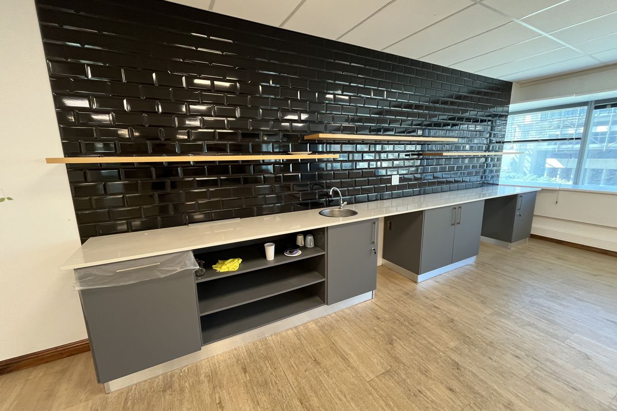 Modern office kitchenette featuring sleek grey cabinetry, a white stone countertop, a stainless steel sink, and a striking black subway tile backsplash.