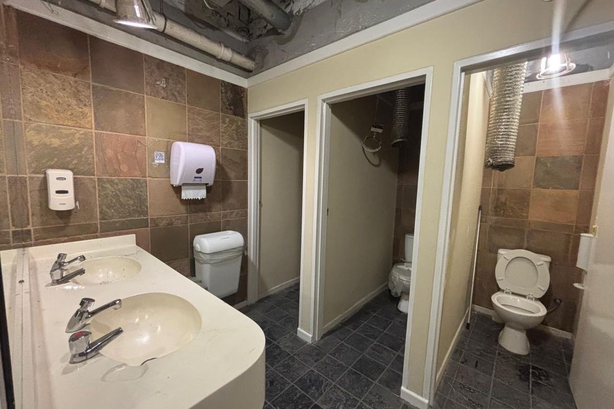 A commercial office restroom featuring a dual-basin vanity, multiple toilet cubicles, and exposed industrial ceiling infrastructure requiring significant renovation.