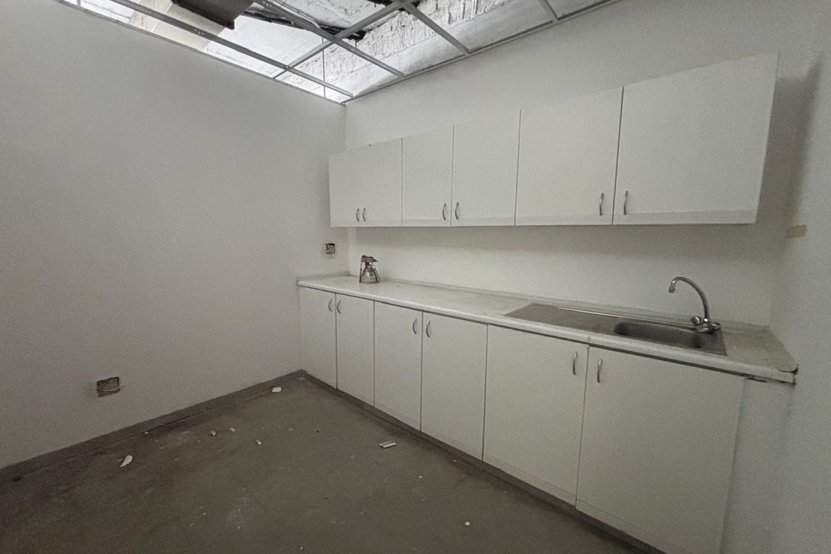 A small office kitchenette featuring white cabinetry, a stainless steel sink, and exposed ceiling grid infrastructure requiring completion.