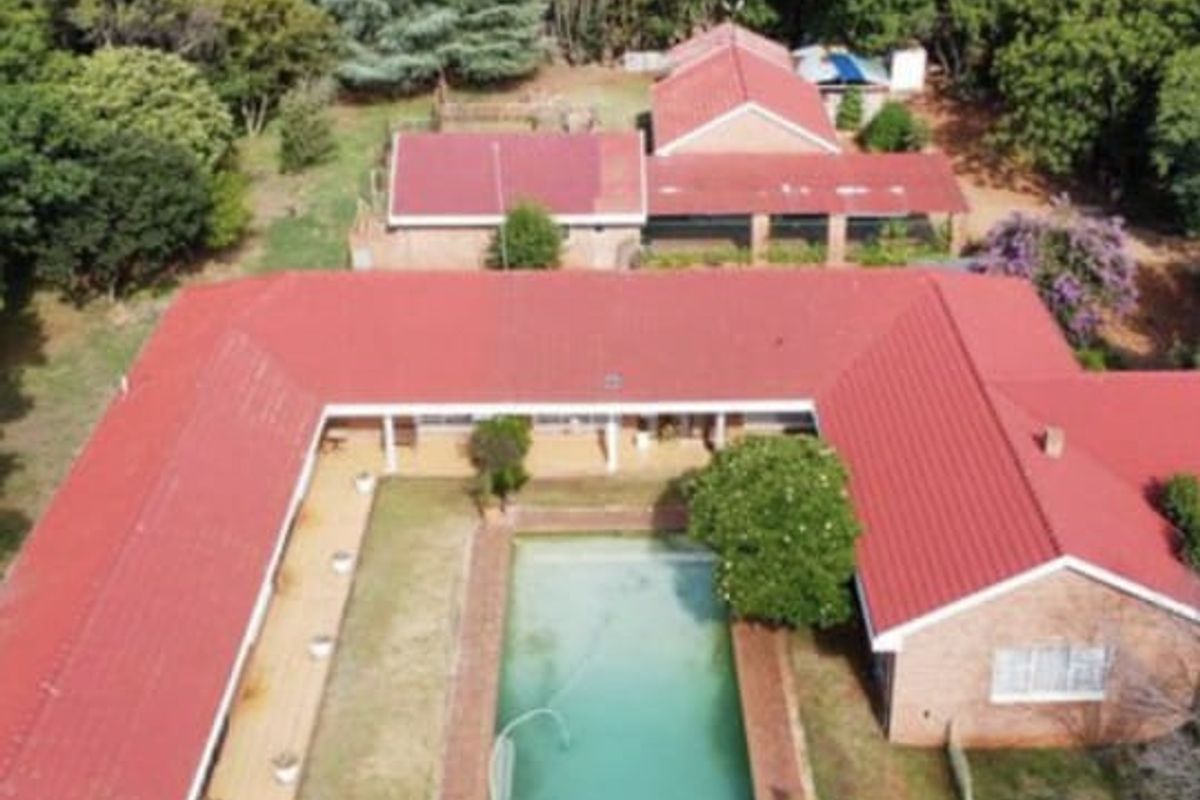 Aerial view of a small holding property featuring a U-shaped dwelling with red metal roofing, a central swimming pool, and surrounding mature garden areas.