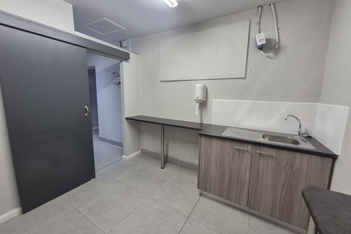 A functional office kitchenette featuring a stainless steel sink, wood-grain cabinetry, tiled flooring, and a wall-mounted soap dispenser.