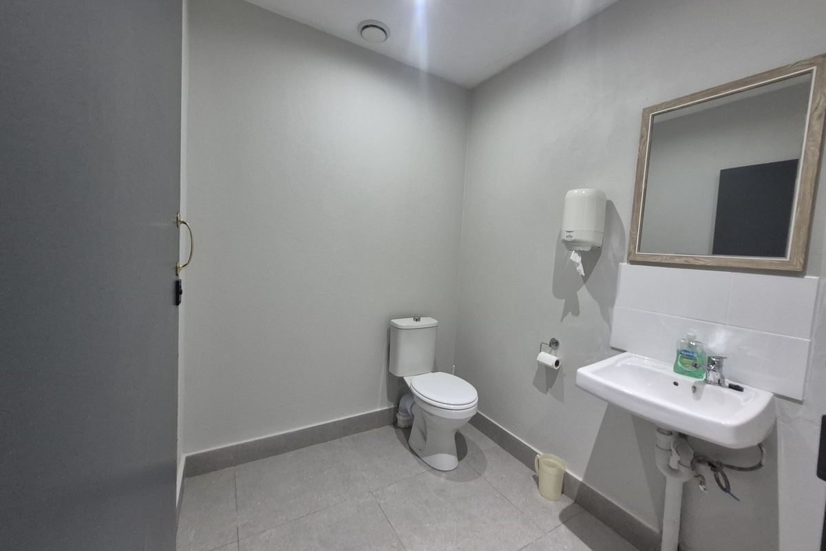A functional office restroom featuring a white ceramic toilet, wall-mounted basin, and a framed mirror.