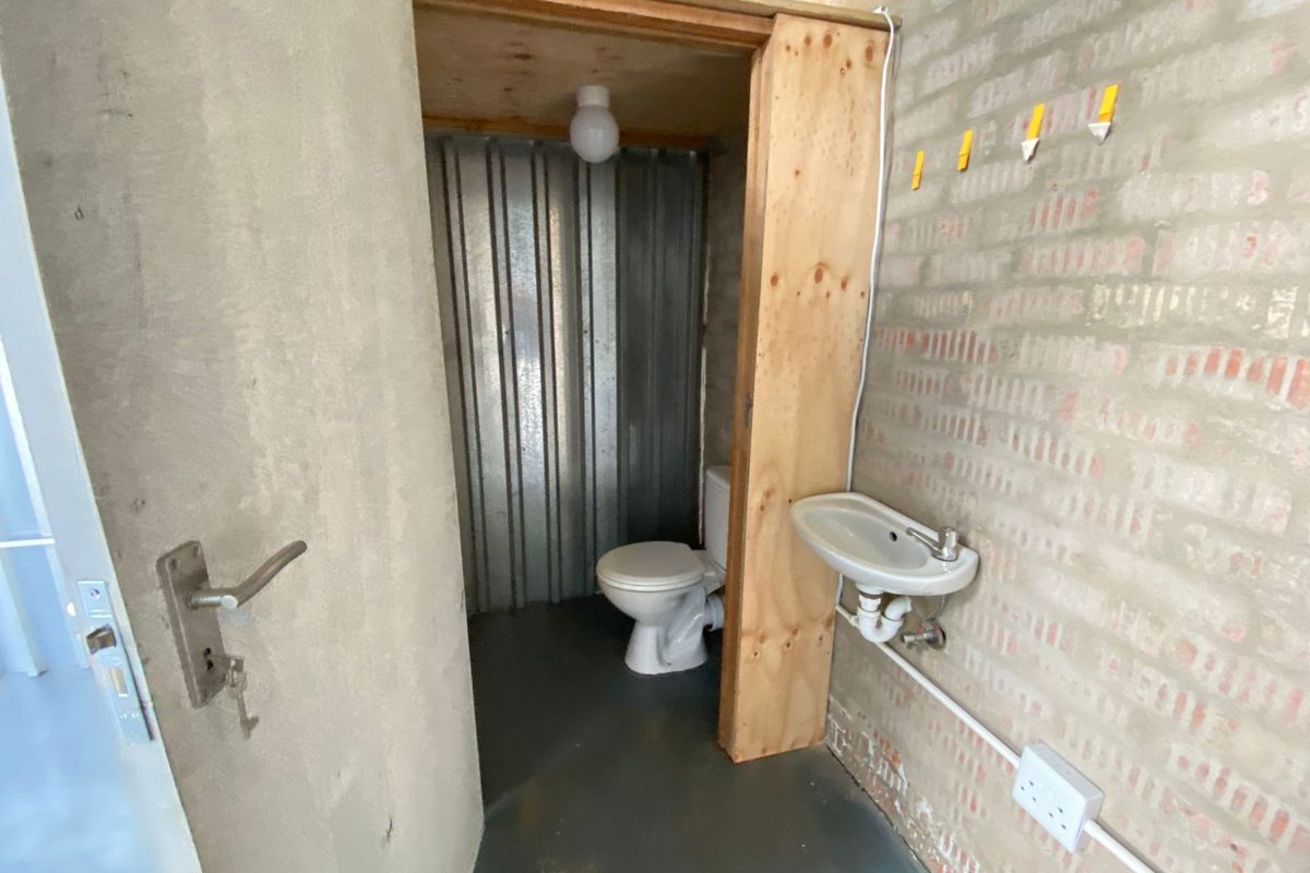 A compact industrial-style bathroom featuring a toilet and wall-mounted basin, enclosed within a plywood structure inside a larger warehouse or workshop space.