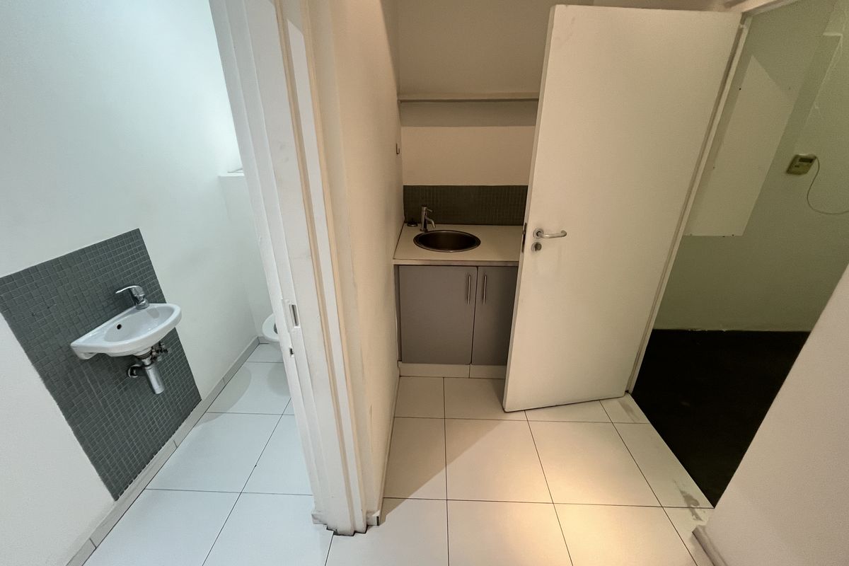A functional office kitchenette area featuring a small sink, built-in cabinetry, and adjacent restroom facilities with tiled flooring.