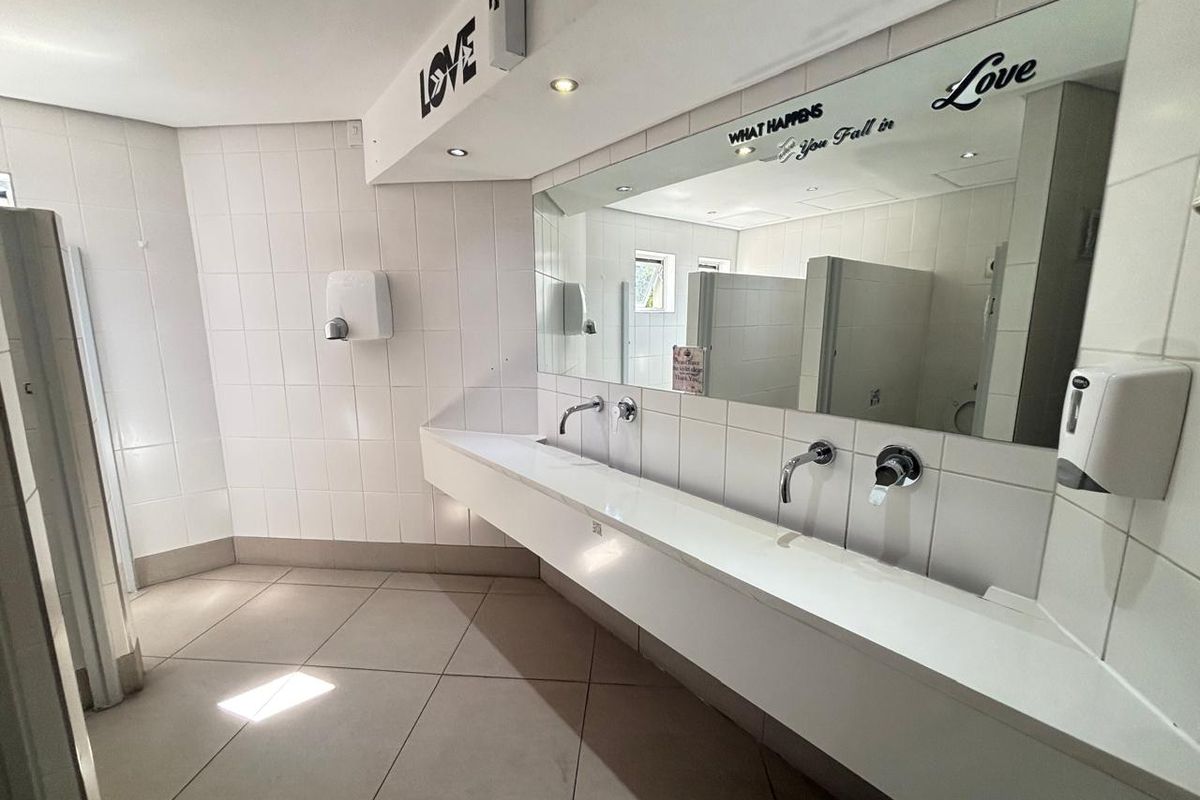 A functional office restroom featuring a long wall-mounted vanity with dual basin faucets, large wall mirror, and tiled flooring.