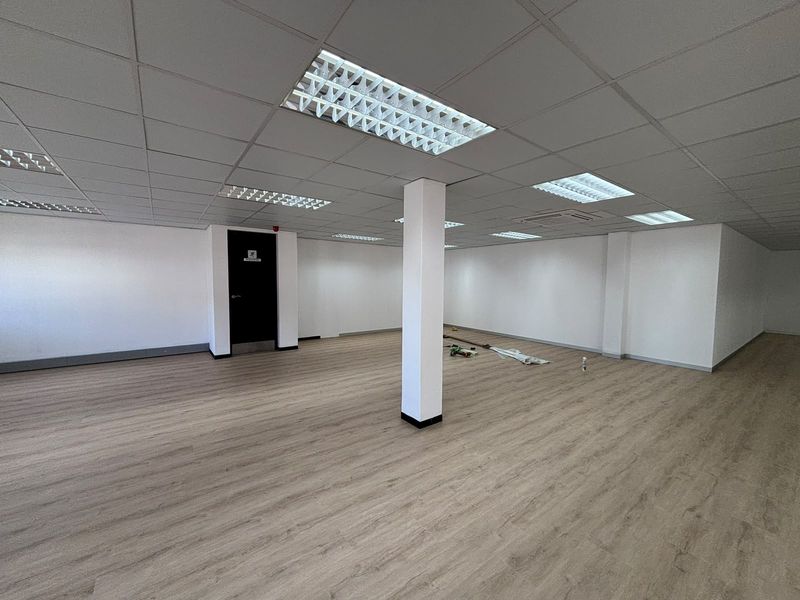 A spacious, open-plan office area featuring modern laminate flooring, a clean suspended ceiling with recessed lighting, and a central structural column.