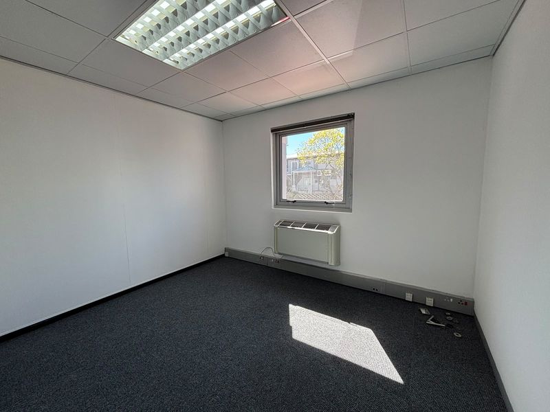 A clean, empty office space featuring grey carpet flooring, a suspended ceiling with recessed lighting, and a wall-mounted HVAC unit.