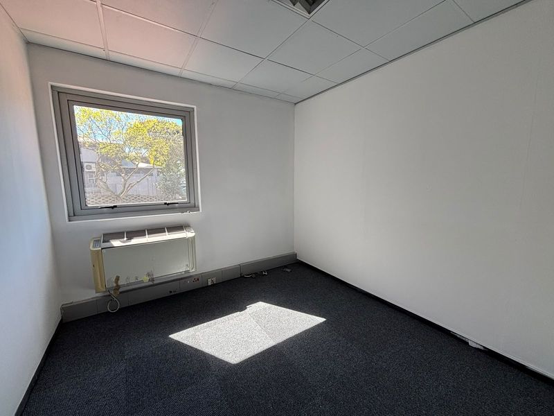 A vacant office space featuring a suspended ceiling, dark carpet flooring, and a wall-mounted air conditioning unit with natural light.