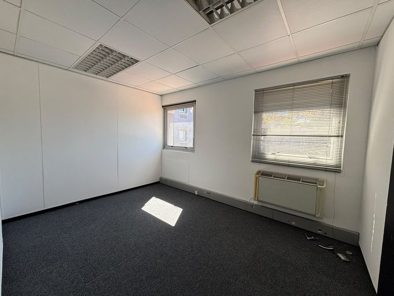 A vacant office space featuring grey carpeting, a suspended ceiling with recessed lighting, and a wall-mounted air conditioning unit.