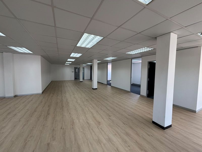 A spacious, open-plan office area featuring modern laminate flooring, a suspended ceiling with recessed lighting, and structural support columns.