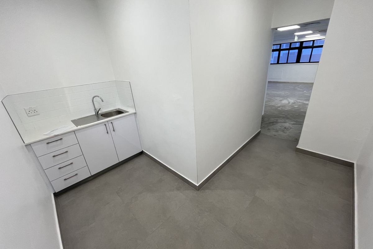 A clean, modern office kitchenette featuring white cabinetry, a stainless steel sink, and tiled splashback, leading into an open-plan workspace with grey flooring.