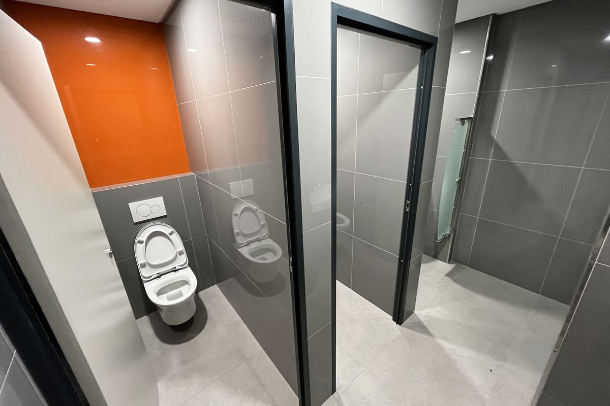 Modern office restroom featuring grey tiled walls, an orange accent wall, and individual toilet cubicles with dark frames.