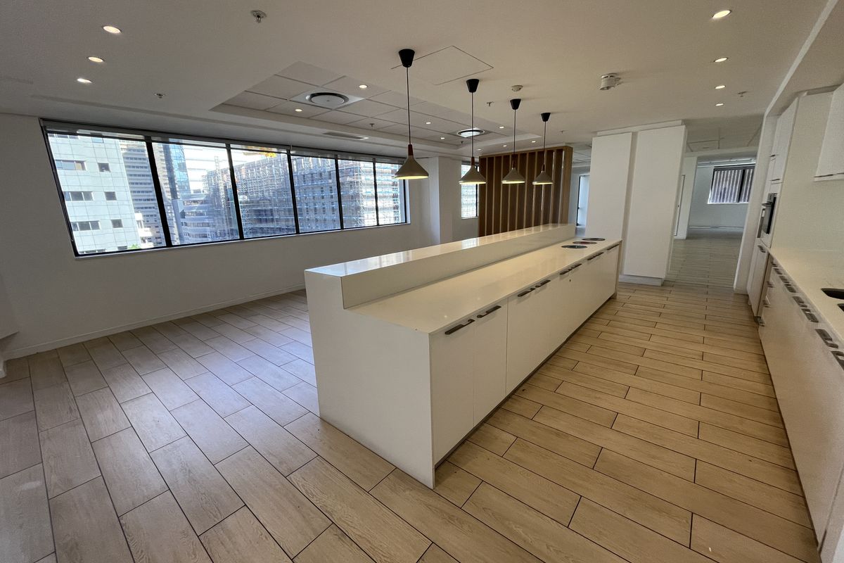 Modern office kitchen area featuring a long central island with white cabinetry, wood-look tile flooring, and large windows providing ample natural light.