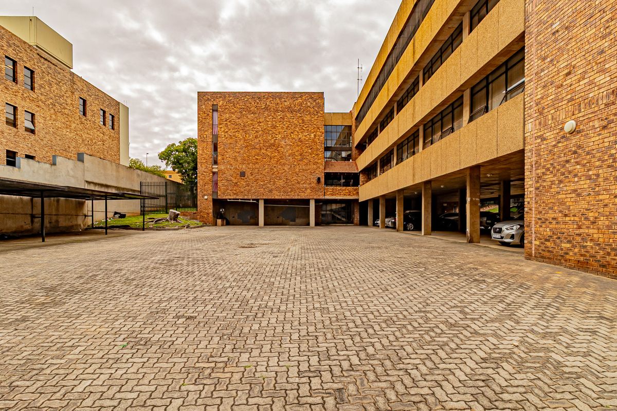Exterior view of a multi-story office building featuring a large paved courtyard and covered parking area with brick facade construction.
