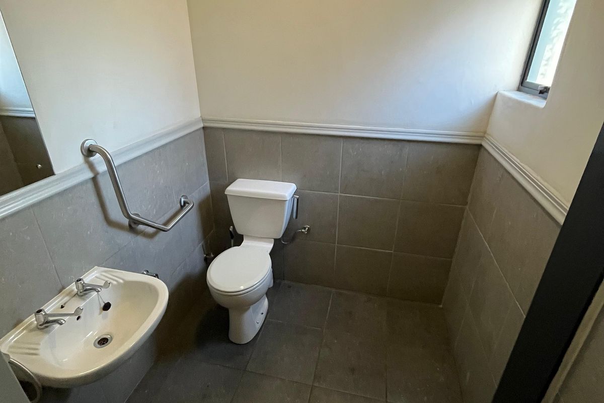 A functional industrial-style restroom featuring a wall-mounted basin, toilet, and safety grab rail with tiled flooring and partial wall tiling.