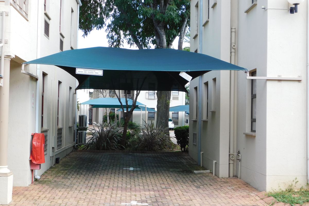 Outdoor parking area featuring a green shade sail structure between two office building wings, with paved ground and established landscaping.