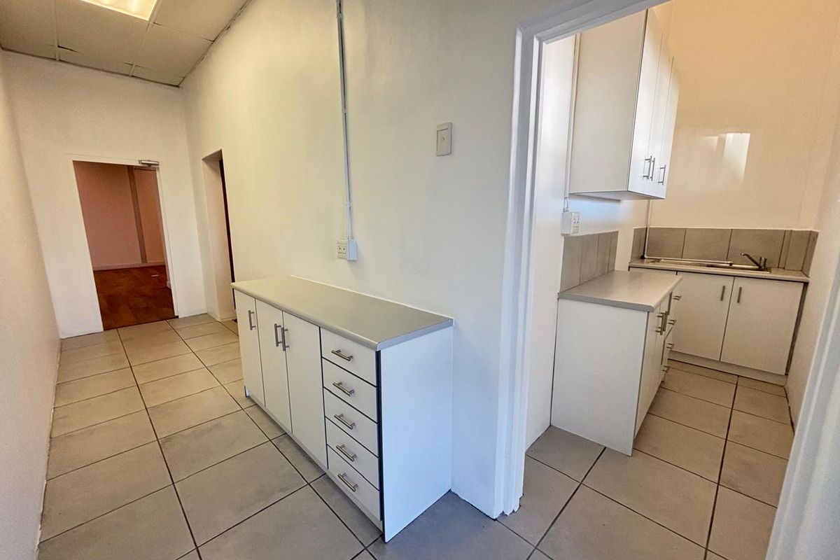 A functional kitchenette area featuring built-in cabinetry, a stainless steel sink, and tiled flooring, with an adjacent hallway leading to further office space.