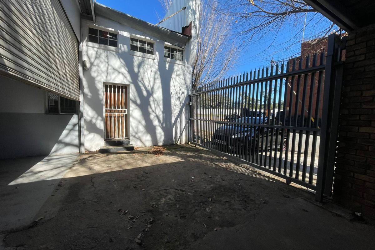 Secure industrial exterior area featuring a metal sliding gate, concrete flooring, and a building facade with security-barred door and windows.