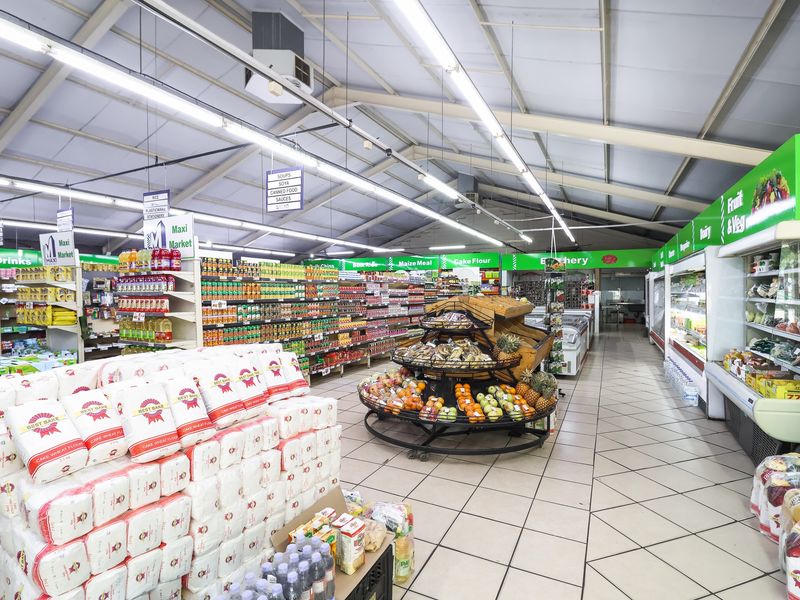 A well-lit retail grocery store interior featuring tiled floors, extensive metal shelving, and refrigerated display units under an exposed steel roof structure.
