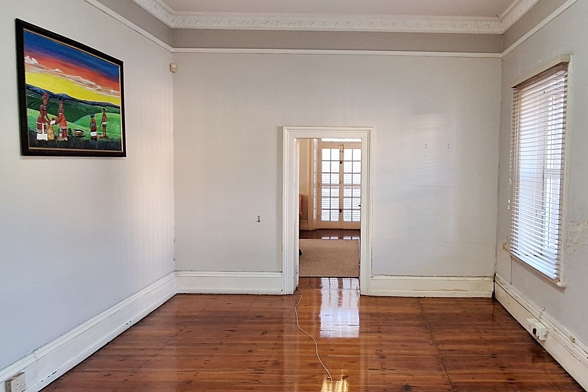An empty living room featuring polished timber flooring, decorative ceiling cornices, and natural light through venetian blinds, leading into an adjacent room.