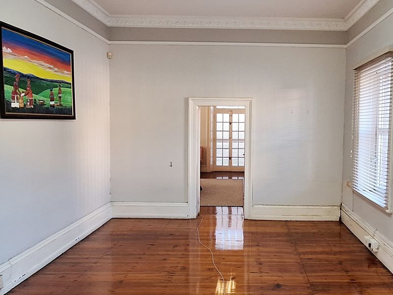 An empty living room featuring polished timber flooring, decorative ceiling cornices, and natural light through venetian blinds, leading into an adjacent room.