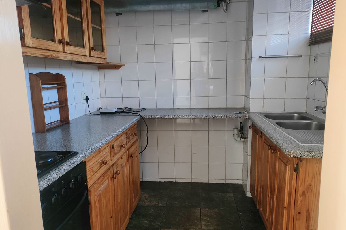 A compact kitchen featuring wooden cabinetry, laminate countertops, a double stainless steel sink, and tiled splashbacks with exposed overhead ventilation ducting.