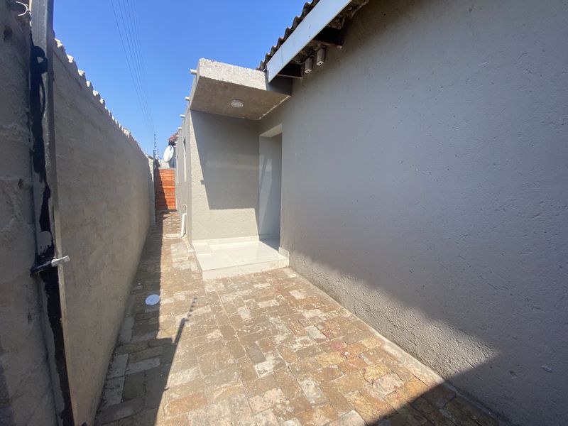 A narrow rear exterior walkway featuring paved flooring, a concrete entrance canopy, and high boundary walls with electric fencing.