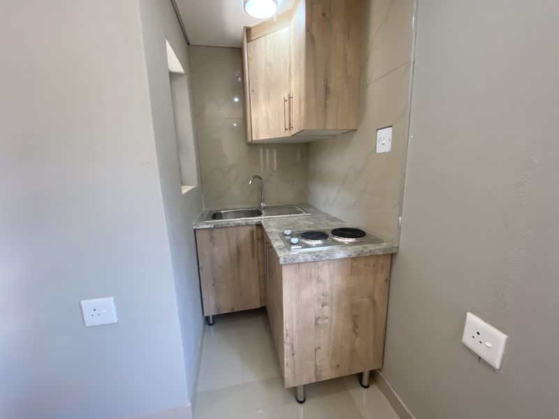 A compact, modern kitchenette featuring wood-finish cabinetry, a stainless steel sink, and a built-in electric hob with tiled splashback.