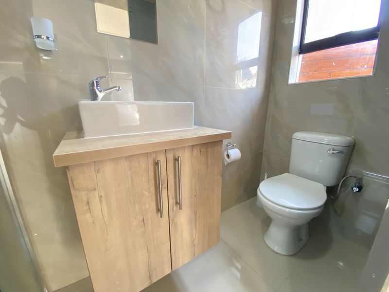 A modern bathroom featuring a wood-finish vanity cabinet with a vessel sink, chrome faucet, and a standard toilet, complemented by neutral wall tiles.