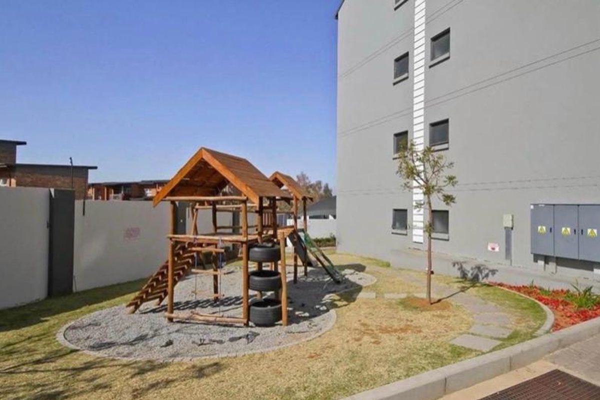 Communal outdoor area featuring a wooden playground structure, paved pathways, and landscaped garden space adjacent to the apartment building.